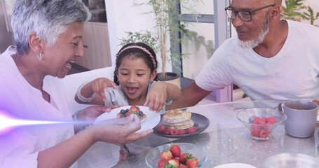 Smiling woman wearing white top offering plate with berries to child at home, pancakes on table