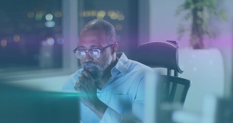 Leaning mature man studying monitors at night office, wearing light shirt and glasses, potted plant