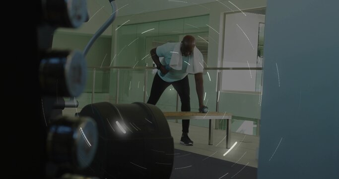 Performing one-arm dumbbell row man wearing light T-shirt, dark pants on bench in gym with towel