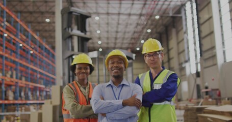 Standing three warehouse workers crossing arms in distribution center, wearing hard hats, vests