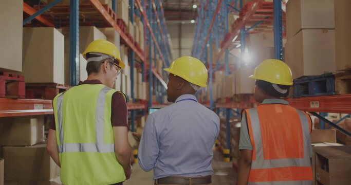Walking 3 warehouse workers wearing yellow hard hats and vests inspecting in warehouse with tablet