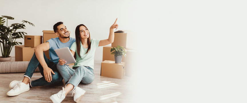 A young couple sits on the floor of their new apartment, surrounded by moving boxes. They are looking forward to decorating their space as they share ideas and laugh together.