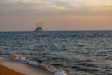 Sunset over Mediterranean Sea with gentle waves on sandy beach and a pirate-themed tourist ship sailing on horizon, illuminated by warm evening light. Antalya, Turkey.

