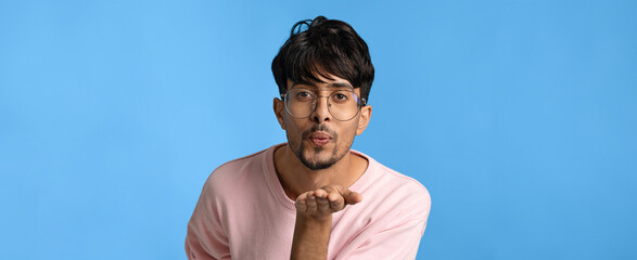 A man in a light pink sweater is blowing a kiss with a playful expression. He is wearing glasses and stands in front of a solid blue backdrop, creating a lighthearted atmosphere.