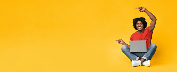 A cheerful young man sits cross-legged on the floor with a laptop in front of him. He smiles and points enthusiastically, showcasing a bright yellow backdrop that adds energy to the scene.