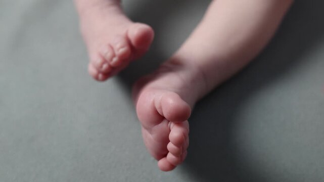 A newborn baby against a gray background. The child is resting with its legs spread out. The mother holds the baby's tiny feet in her hands. The girl strokes the doll's toes