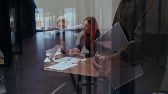 Two male professionals debate business strategy and charts while coworkers continue working behind them. Shot through the reflective window surface creating a modern corporate narrative
