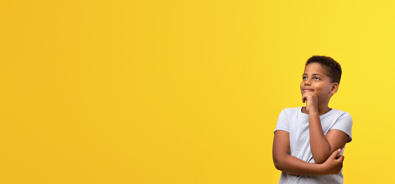 A young boy with short hair is standing against a vivid yellow wall. He looks thoughtful, resting his chin on his hand with a smile. The bright color enhances his calm demeanor.