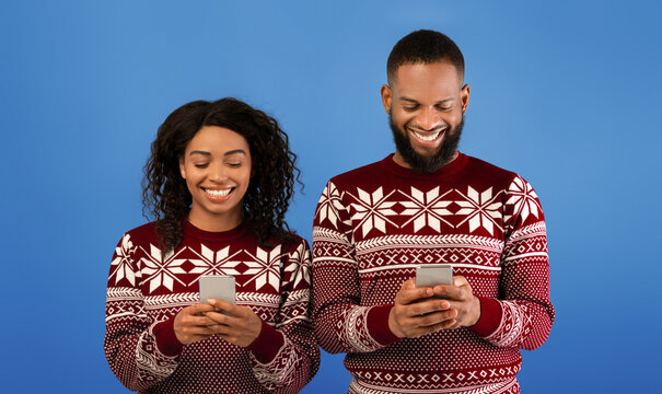 Joyful black couple in Christmas sweaters playing together on smartphones, standing over blue studio background. Happy african american spouses using social media