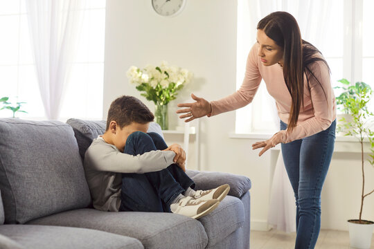 Family conflict. Angry emotional mother scolding upset preteen boy sitting on sofa with arms around knees. Sad child avoiding eye contact while experiencing stress during serious conversation at home.