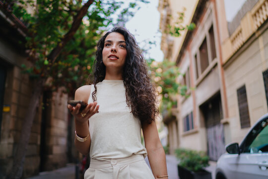 Curious young woman holds smartphone, interacting via voice command, showcasing seamless AI integration and smart technology in natural urban use.