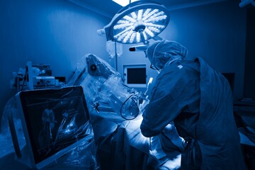 A surgeon, wearing full protective gear, performs a knee replacement using robotic-assisted technology in a sterile, blue-lit operating room at a hospital © Damian