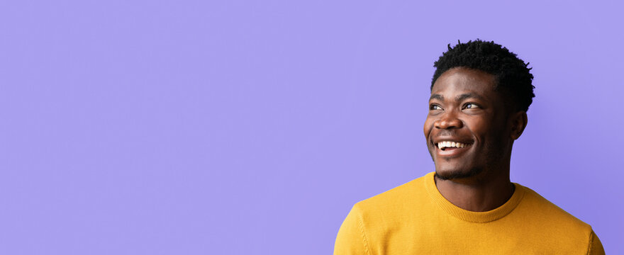 A man grins joyfully while looking to his right. He wears a bright yellow sweater, exuding warmth and positivity, with a simple purple backdrop highlighting his cheerful demeanor.