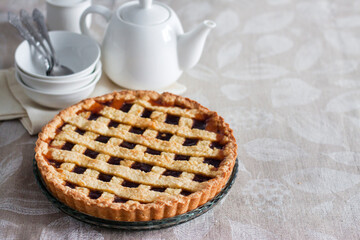 Freshly baked lattice pie on a rustic table beside stacked plates, bowls, and a white teapot, creating a cozy homemade dessert scene.