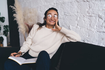 Smiling freelancer relaxes during creative session with notebook in hand, enjoying digital freedom and balancing analog planning with online ambitions in cozy home workspace setup.