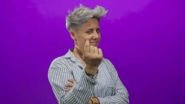 Woman in striped shirt and watch beckons with finger in purple studio while smirking at camera; playful invitation.