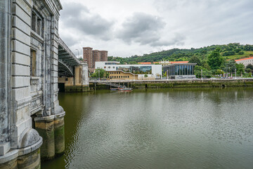 View of the embankment from the river in the city of Bilbao, Spain. A large stone bridge is visible on the left side The photo was taken in cloudy weather, with the sky covered in clouds