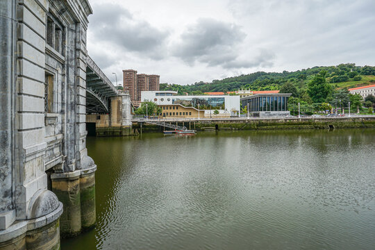 View of the embankment from the river in the city of Bilbao, Spain. A large stone bridge is visible on the left side The photo was taken in cloudy weather, with the sky covered in clouds