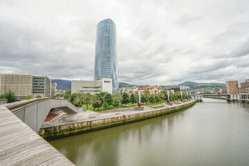 A view of the embankment in the city of Bilbao, Spain, from the Nervion River A large, tall, blue skyscraper is located on the embankment. The weather is cloudy, with an overcast sky
