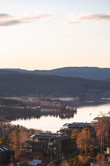 Evening light reflects over calm Norwegian lake near Oslo, mirroring dark pine forests and distant houses in tranquil Nordic autumn atmosphere