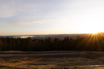 Golden rays touch treetops above Oslo as sun sets over hills and fjord, creating a warm balance between Nordic wilderness and urban horizon. Grefsenkollen