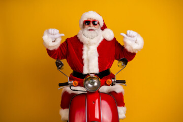 Santa Claus on a red vintage motorcycle poses in a bright yellow studio with thumbs up for Christmas celebration and festive joy