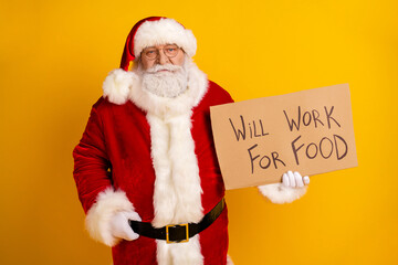 Santa Claus in a red and white suit holds a cardboard sign reading will work for food against a bright yellow background