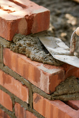 Close-up of bricklaying with fresh mortar and a trowel applying cement between red bricks. The scene highlights construction work, masonry craft and the detailed process of building a brick wall.