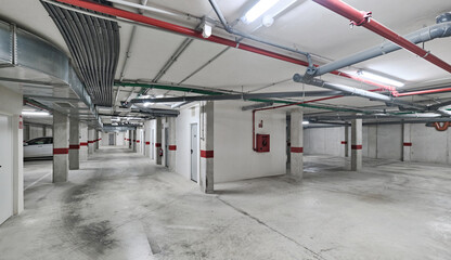Clean and spacious underground parking garage with ventilation ducts, pipes, and red safety markings on concrete walls and columns