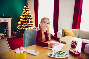 Funny cozy Christmas scene with a smiling girl drawing at a colorful table by a lit Christmas tree in a warm home