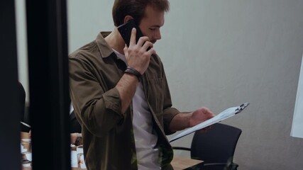 A serious businessman holds documents and talks on the phone while leaning on a wooden table inside a modern office. His colleagues are visible in the background, working and discussing business tasks - Powered by Adobe