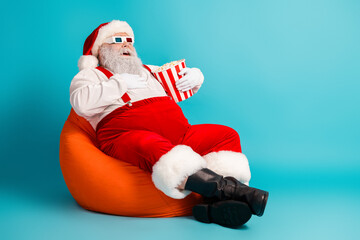 Santa in red suit on orange beanbag laughing with popcorn bucket under a blue background for a...