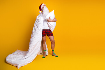 Funny santa in pajama holds pillow and blanket in a bright yellow studio creating a cozy Christmas vibe for home life and holiday fun