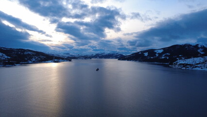 A dramatic drone view of a fjord with a ship in the center, encircled by towering snowy mountains under a deep blue sky filled with clouds. This serene scene captures adventure and exploration. 