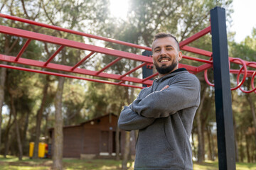 Smiling man standing with crossed arms in outdoor gym, enjoying calisthenics workout and modern fitness lifestyle