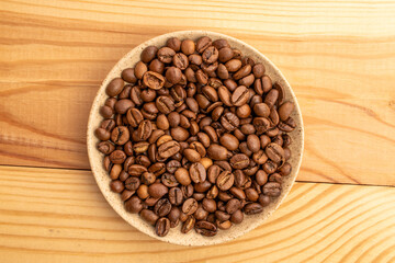 Roasted coffee beans in a ceramic saucer on a wooden table, top view.