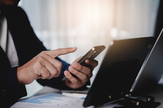 A man is using his cell phone while sitting at a desk with a laptop and papers
