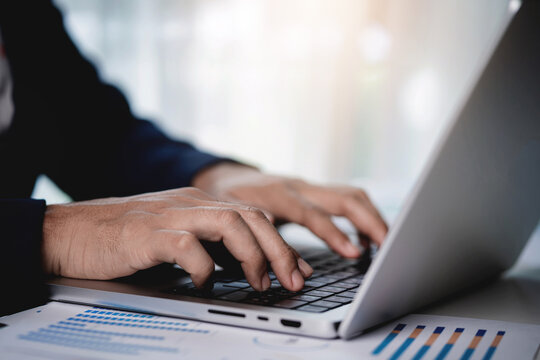 A person is typing on a laptop computer with a stack of papers in front of them