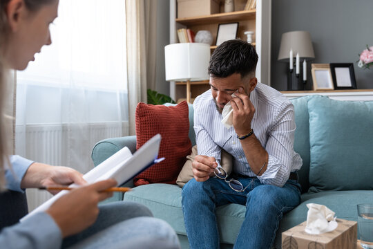 Business professional in psychotherapy at home: man shares feelings with therapist; tissues and water nearby, daylight and decor suggest a confidential counseling session focused on mental wellbeing