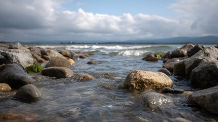 Gentle waves wash over smooth wet stones on a rocky shore with a vast cloudy sky stretching towards the horizon