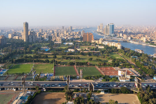 View of vibrant green spaces and buildings nestled along the Nile, with a bustling highway below, all under a hazy sky, Zamalek, Cairo Governorate, Egypt.
