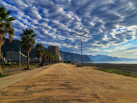 A wide, empty concrete promenade runs along the Black Sea coast in Gonio, Georgia, lined with palm trees and modern high-rise buildings in the distance, capturing an off-season mood.