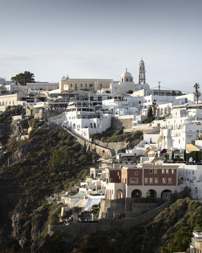 View of whitewashed buildings clinging to the cliffside, illuminated by the sun, creating a striking contrast against the dark volcanic rock, Fira, Thira, Greece.