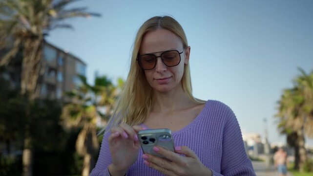 Young blonde woman in sunglasses use smartphone for texting and browsing social media while walking outdoors in city park with palm trees on bright and sunny day