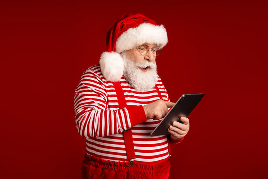 Santa uses a tablet in a red festive setting wearing striped shirt suspenders and hat as he smiles and checks holiday messages for Christmas advertising