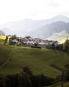 View of a cluster of houses nestling in the verdant hills, with soft light casting shadows and the mountain range fading into the horizon, Slovenia.