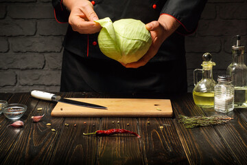 A chef holds a fresh cabbage in a kitchen, with a wooden cutting board, knife, and various ingredients nearby. The warm light creates a welcoming cooking environment for meal preparation