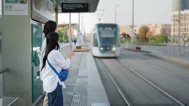 Asian tourist couple waiting for tram at city station outdoors. Public transport lifestyle in Barcelona