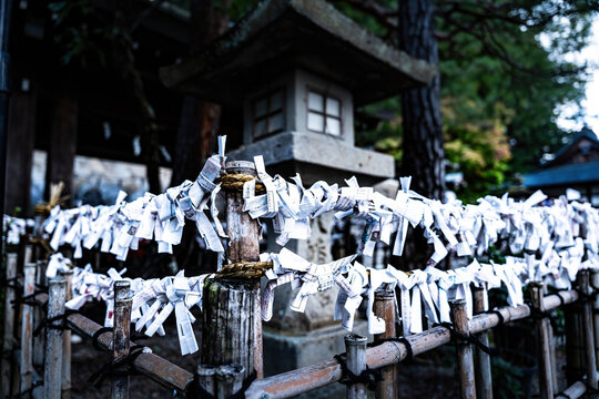 View of weathered wooden fence adorned with countless white paper fortunes fluttering gently, a stone lantern standing guard in the background, Takayama, Gifu, Japan.