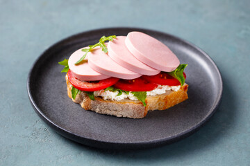 Fresh open sandwich with boiled sausage, tomato slices, cottage cheese and arugula on rustic bread. Grey background. Close up.